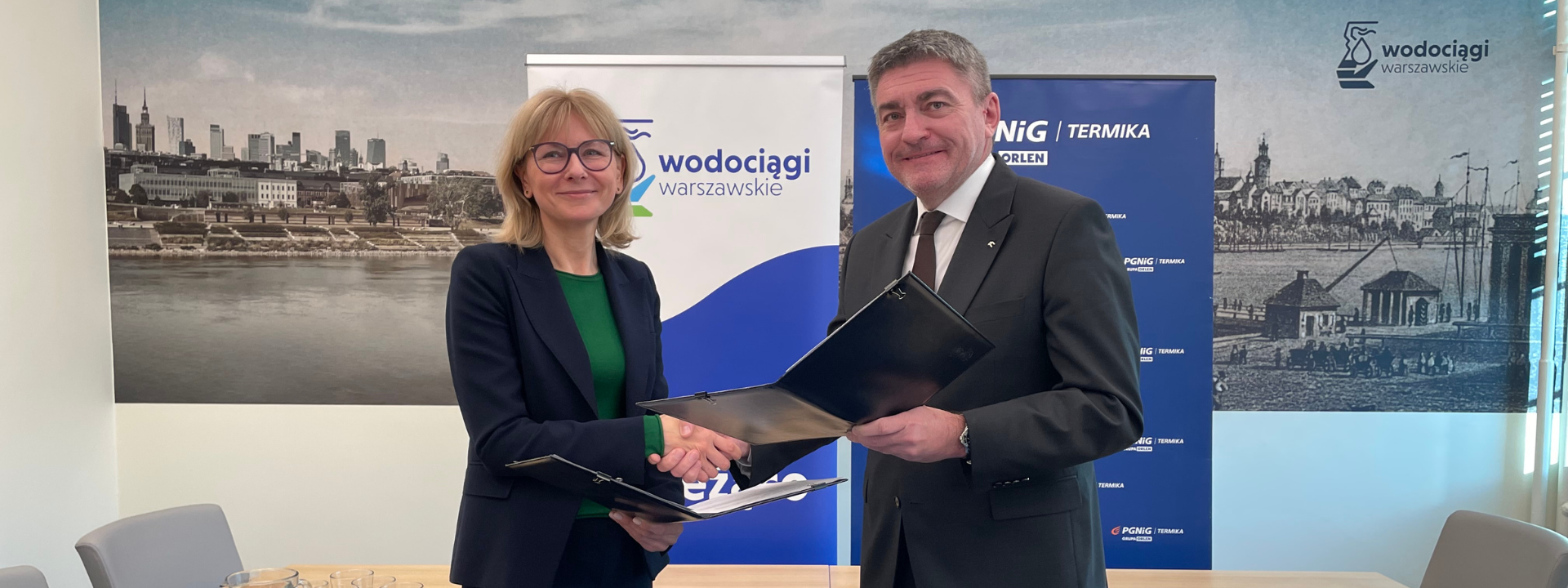 Prezes Wodociągów Warszawskich i prezes PGNiG Termika podają sobie ręce i przekazują dokumenty w teczce. W tle tapeta z panorama Warszawy.