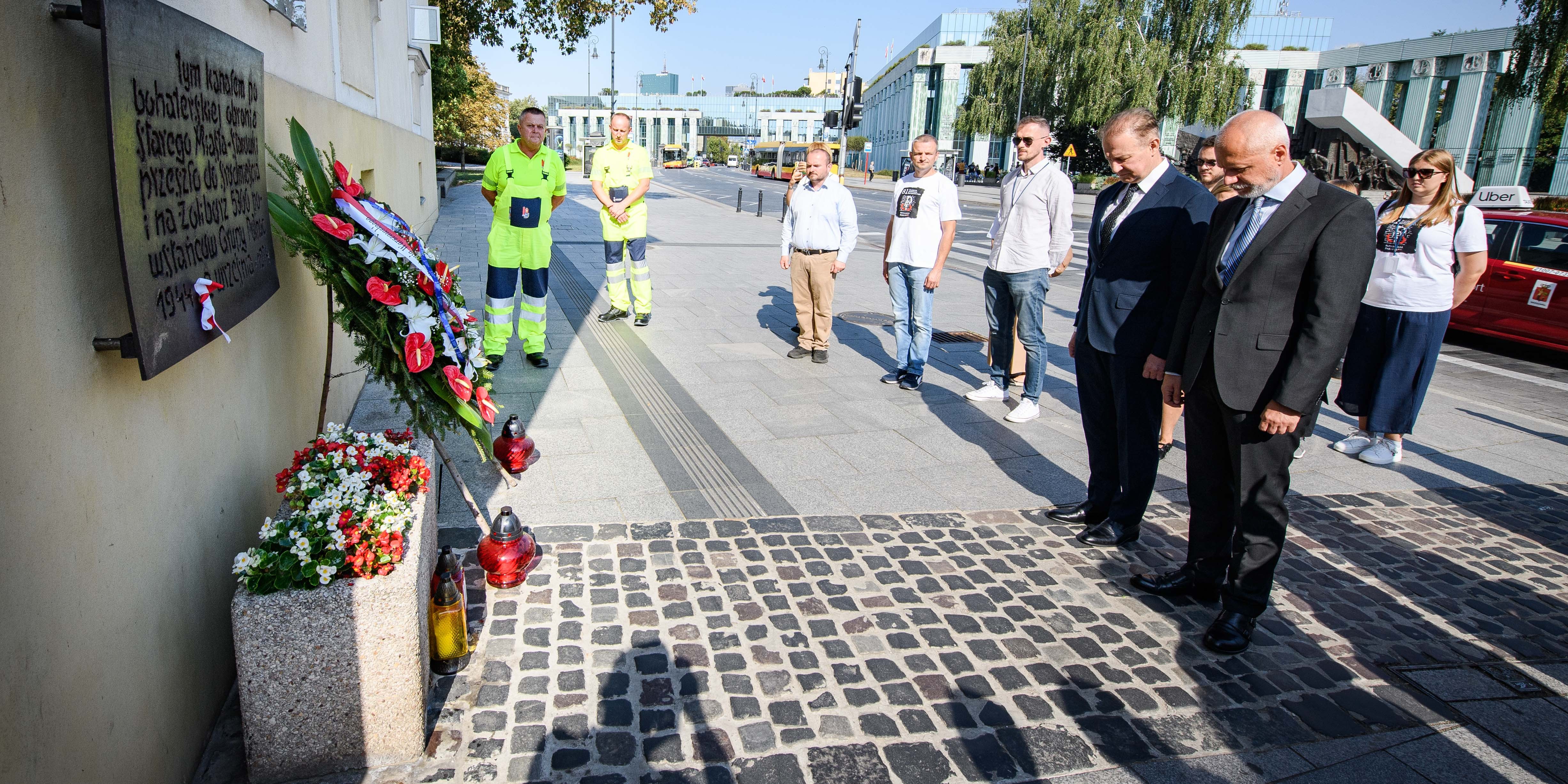 Grupa osób stojących przed tablicą zawieszoną na ścianie budynku. Pod murem stoi wieniec.
