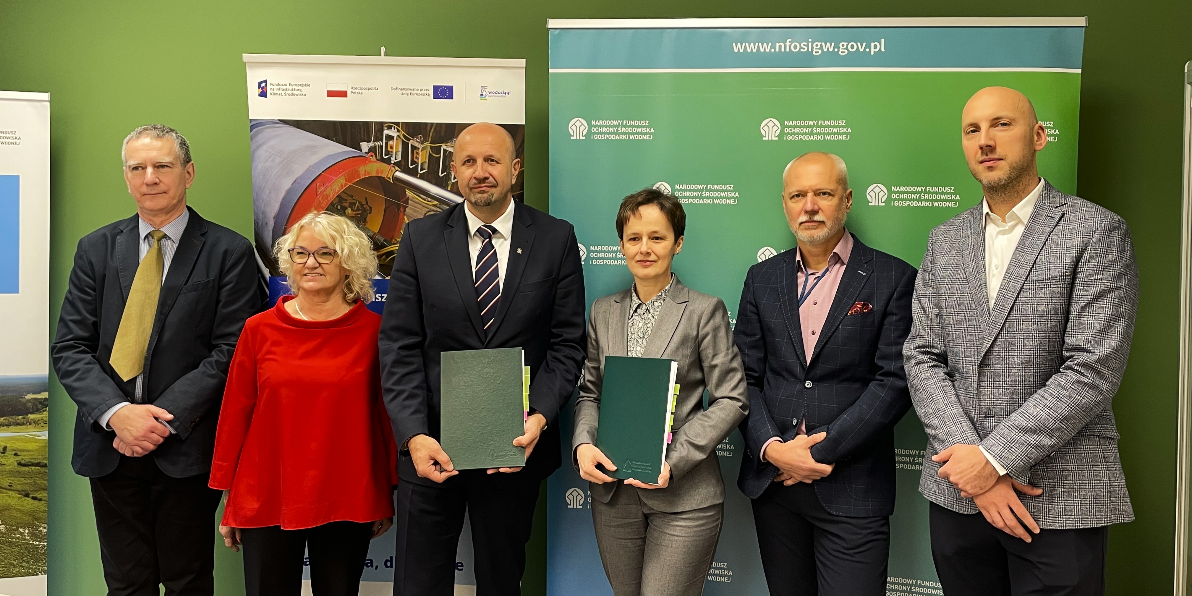 Przedstawiciele Wodociągow Warszawskich oraz Narodowego Funduszu Ochrony Środowiska i Gospodarki Wodnej stojący w rzędzie, dwoje z nich trzyma teczki z dokumentami, na tle plansz z logotypami unijnymi